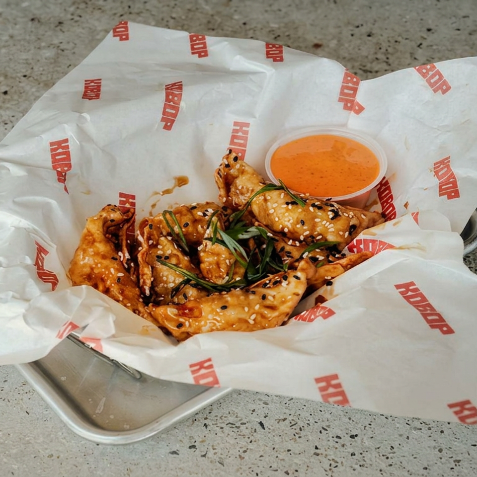 Crispy sesame-coated dumplings with herbs and orange dipping sauce served on branded paper in a metal tray.