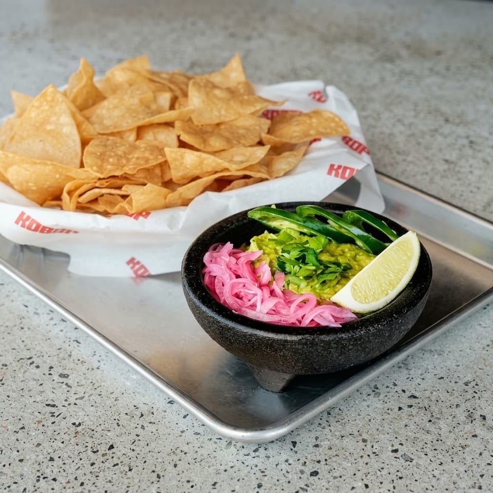 Crispy tortilla chips accompany guacamole topped with pickled onions, jalapeños, cilantro, and a lime wedge on a restaurant tray.
